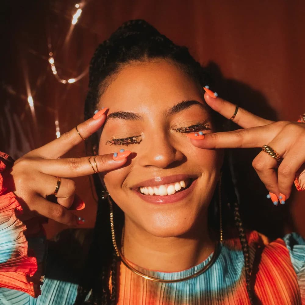 Tiana, the artist behind Disco Tips, smiling with her eyes closed and peace signs framing her face, wearing glitter lashes and coral nails with blue dots.