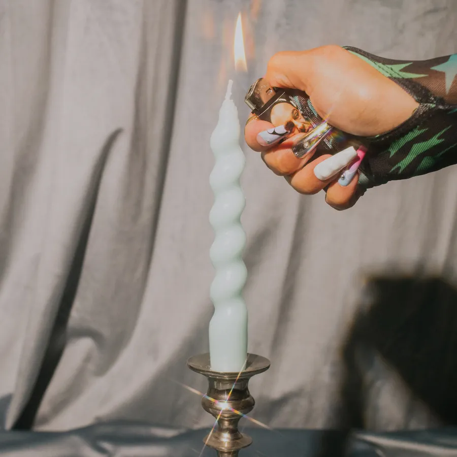 A hand in long nails with pastel and chrome art lighting a pale mint twisted candle in a brass candlestick.